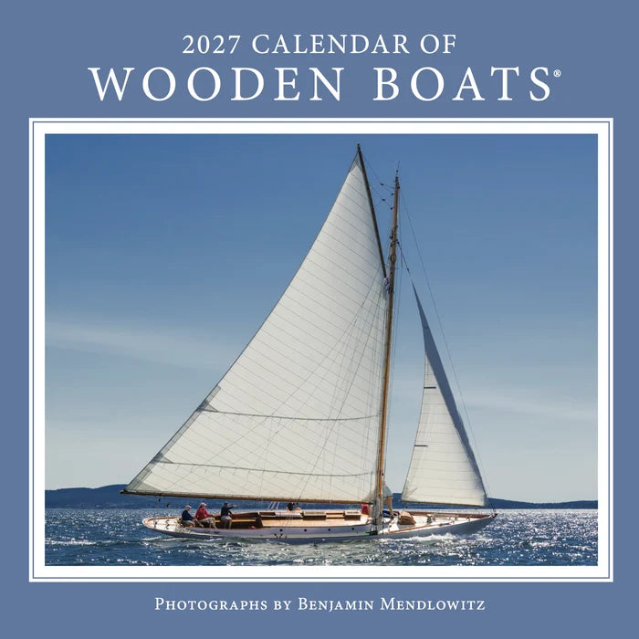 A classic wooden sailboat with white sails glides across tranquil blue waters beneath a cloudless sky, capturing the enduring allure of maritime tradition. The cover features the title “2027 Calendar of Wooden Boats,” with photography by Benjamin Mendlowitz.