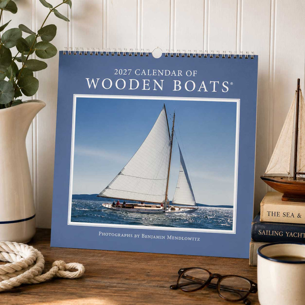 The 2027 Calendar of Wooden Boats, featuring exquisite nautical craftsmanship and a stunning sailboat cover photo, is displayed on a table next to books, a cup, a plant, glasses, and a small model sailboat.