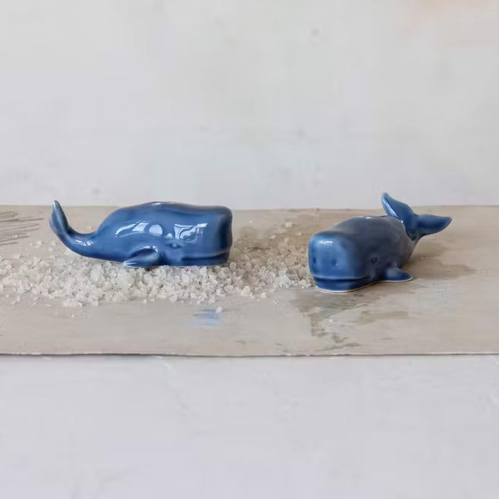 The Stoneware Whale Salt & Pepper Shakers, in a small blue whale design, sit on a wooden surface with coarse salt nearby, perfect for adding nautical flair to your kitchen decor.