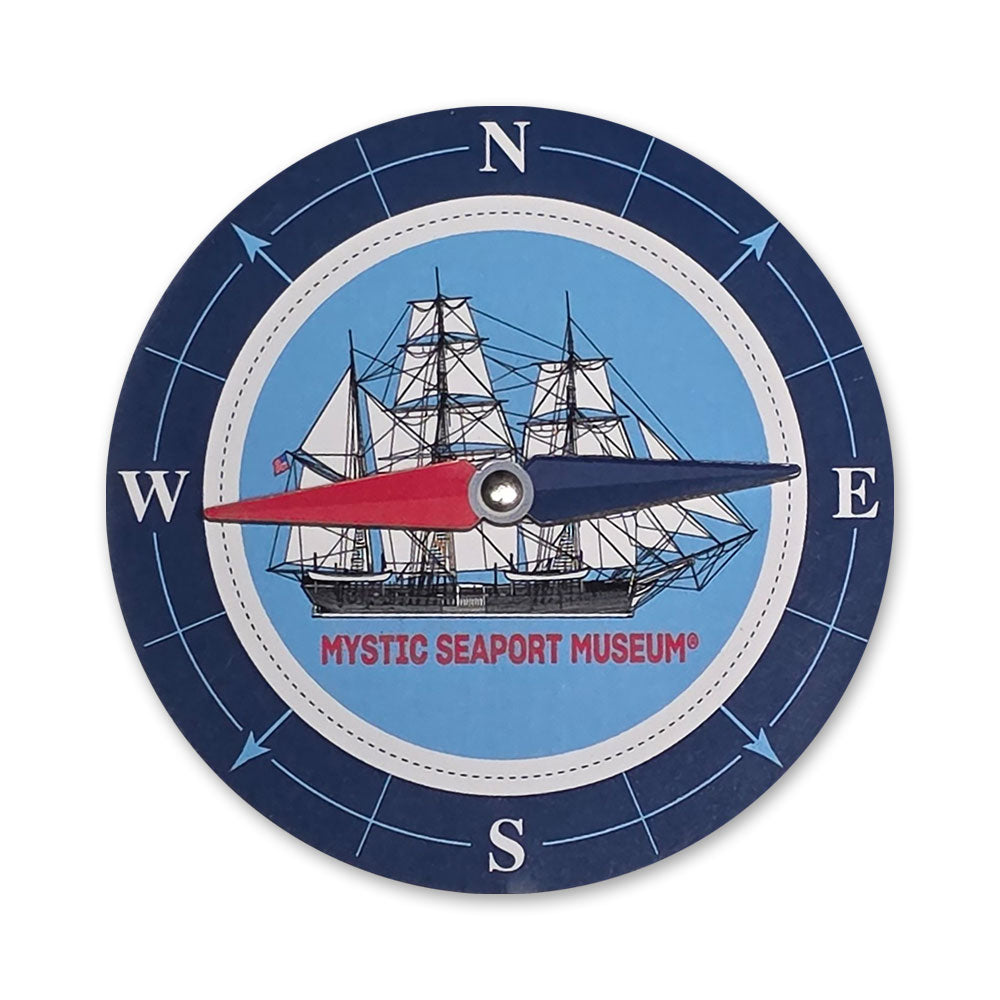 The Mystic Seaport Museum Tall Ship Spinner Magnet features a nautical compass rose with a central ship illustration and the museum’s name. Red and blue needles point west and east, making it ideal for maritime heritage collections.
