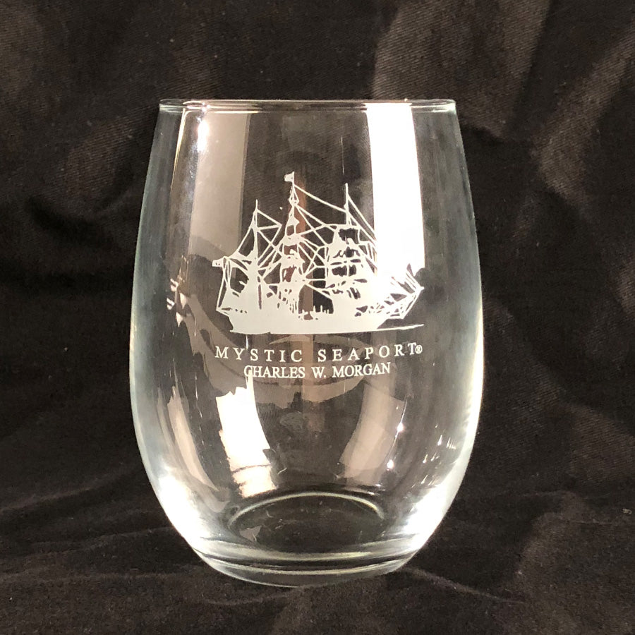 The CHARLES W. MORGAN Stemless Wine Glass features a white-etched sailing ship and Mystic Seaport® Charles W. Morgan text on clear glass, crafted by Susquehanna Glass, shown here against a black background.